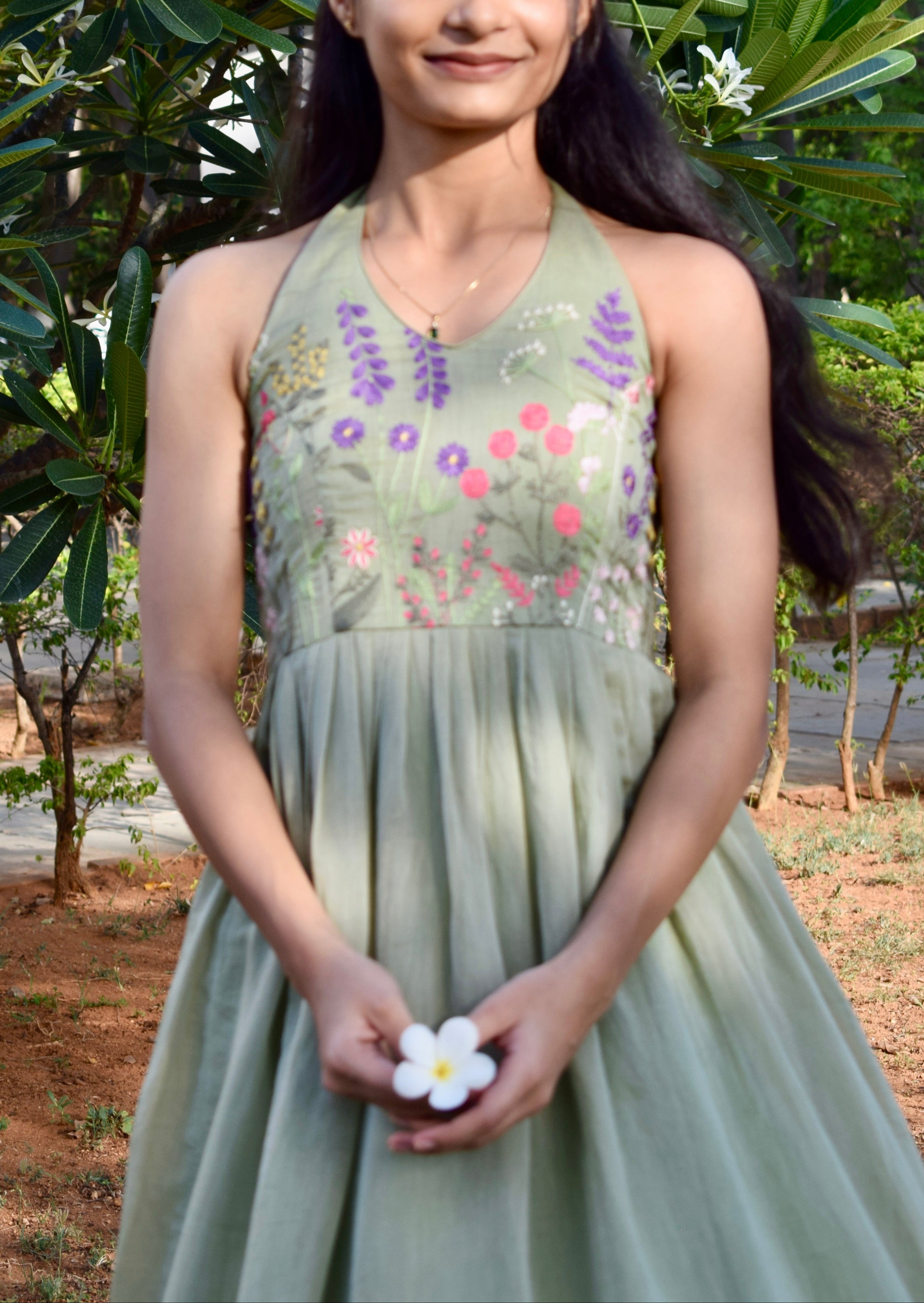 Woman in a green dress with floral embroidery standing outdoors among trees and plants.