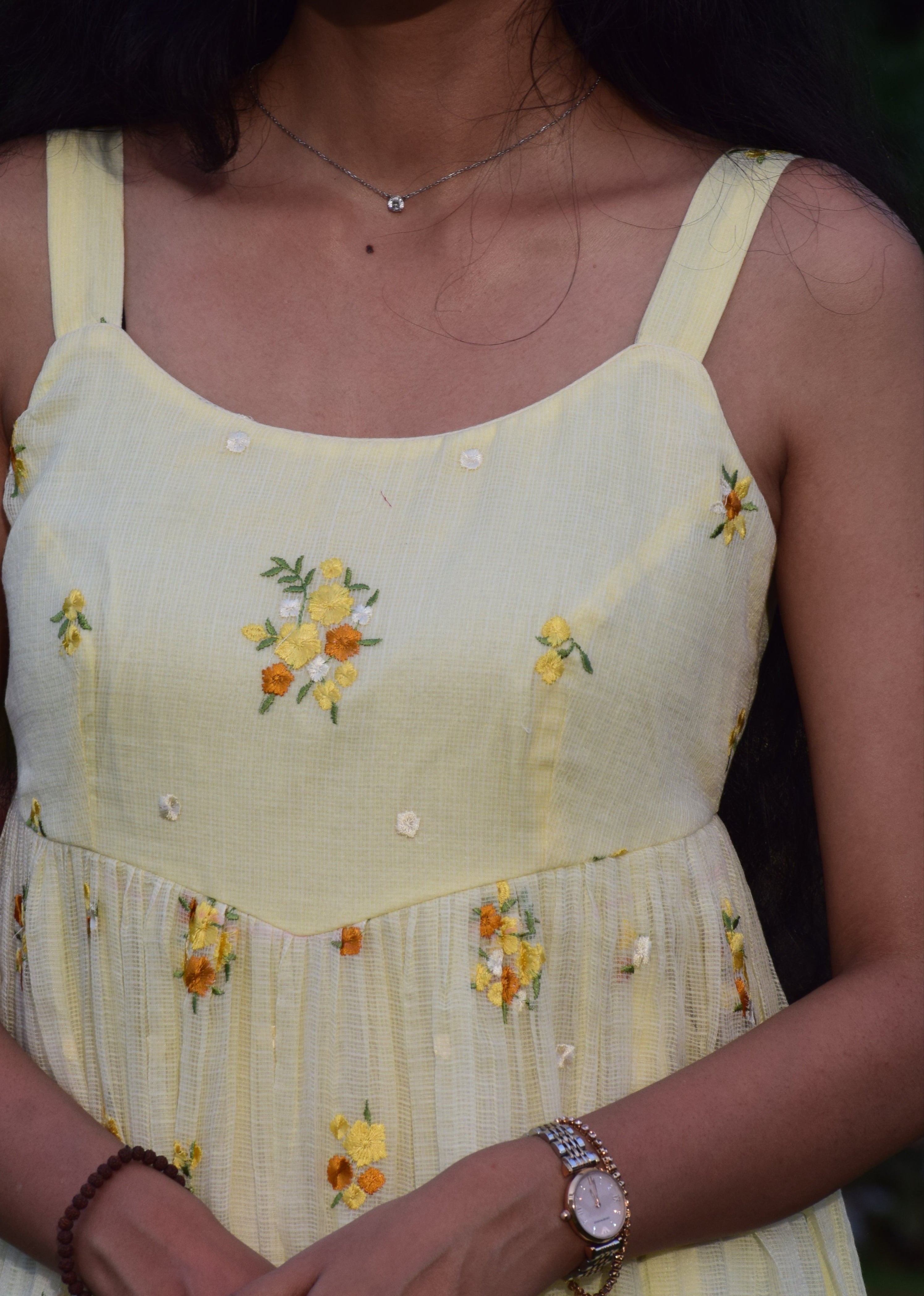 Woman wearing a yellow floral dress with her arms crossed, surrounded by greenery.