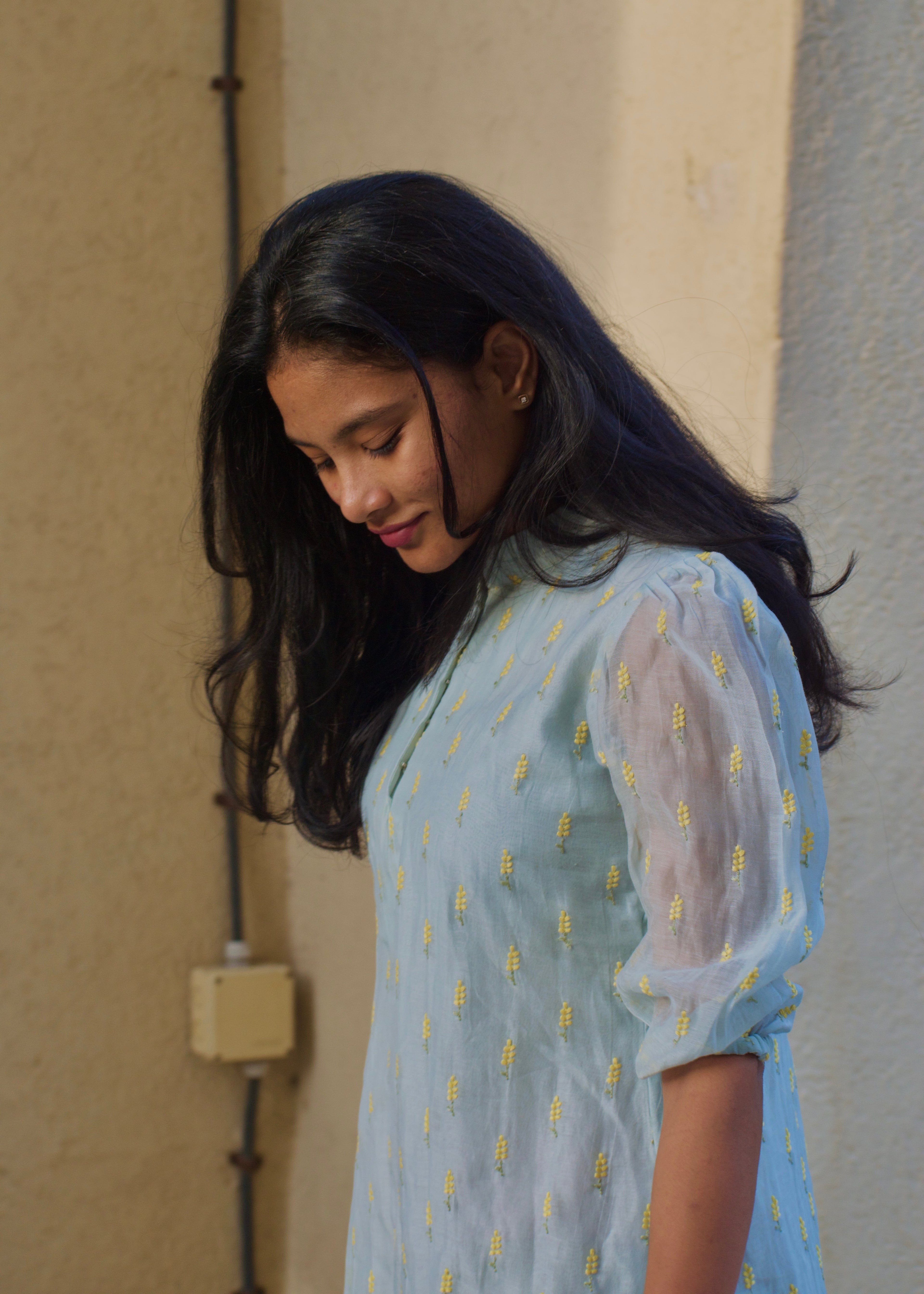 Woman wearing a light blue dress with yellow patterns, standing against a neutral wall.