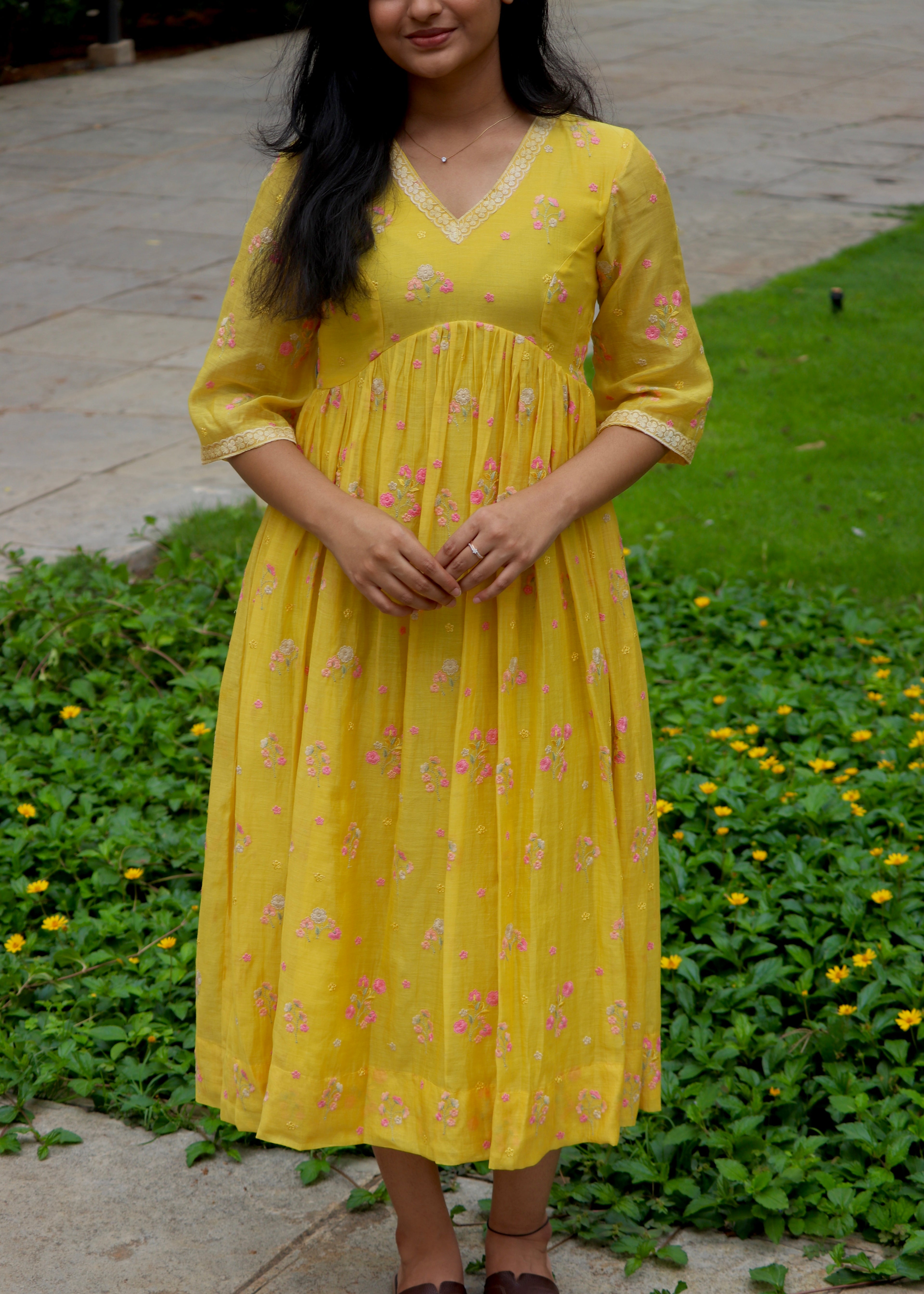 Woman wearing a yellow dress with floral patterns standing outdoors on a grassy area.