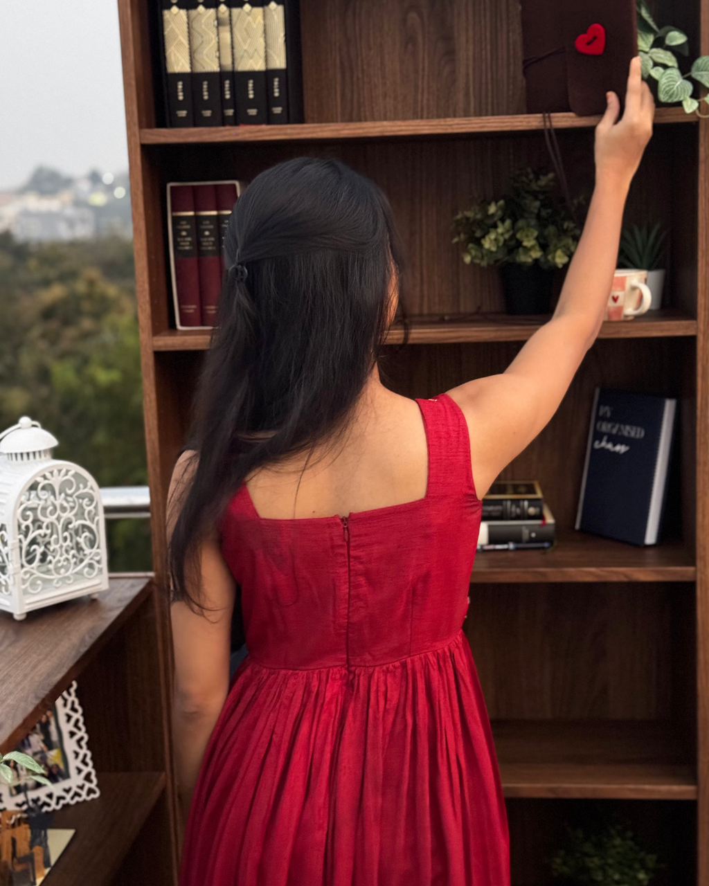 Woman in a red dress reaching up to a wooden bookshelf.