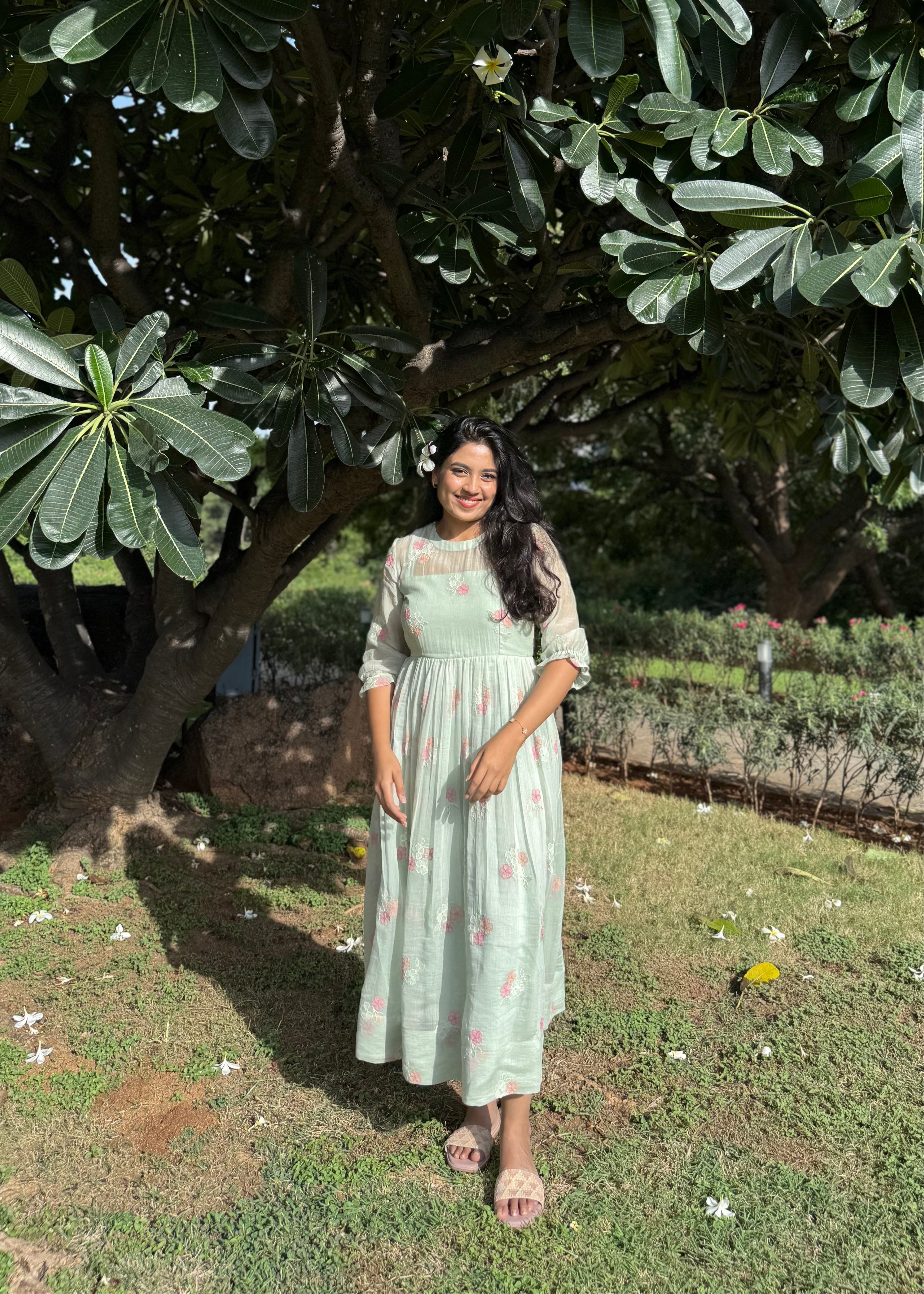 Woman in a light green dress standing under a large tree with greenery around.