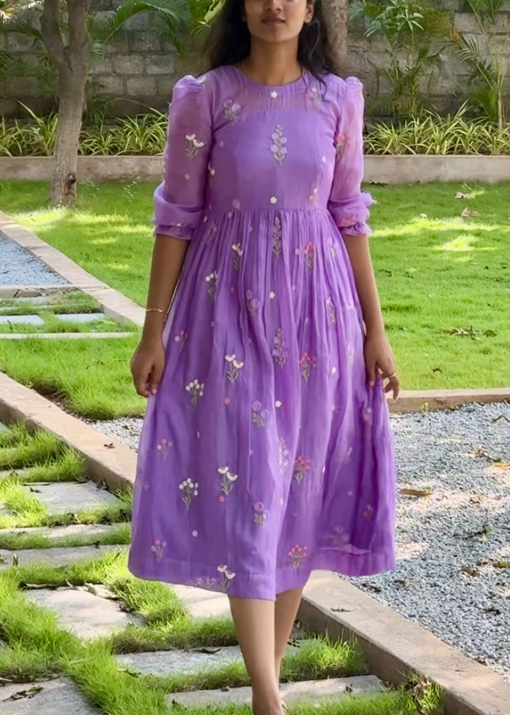 Woman wearing a purple dress with floral patterns standing outdoors on a grassy area.