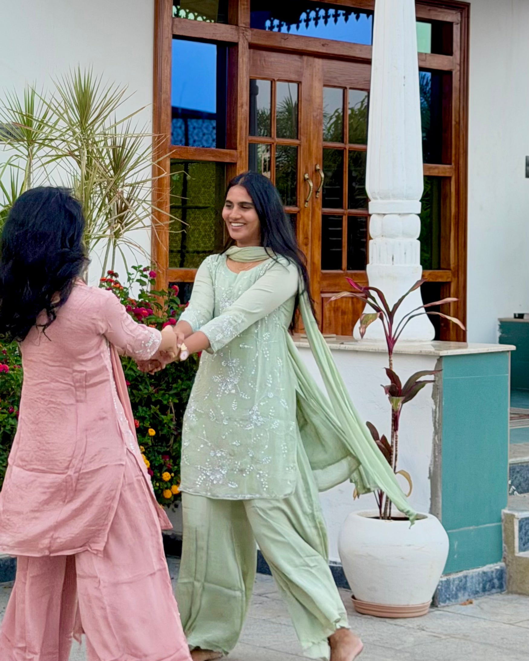 Two women shaking hands outside a building with plants and flowers in the background.