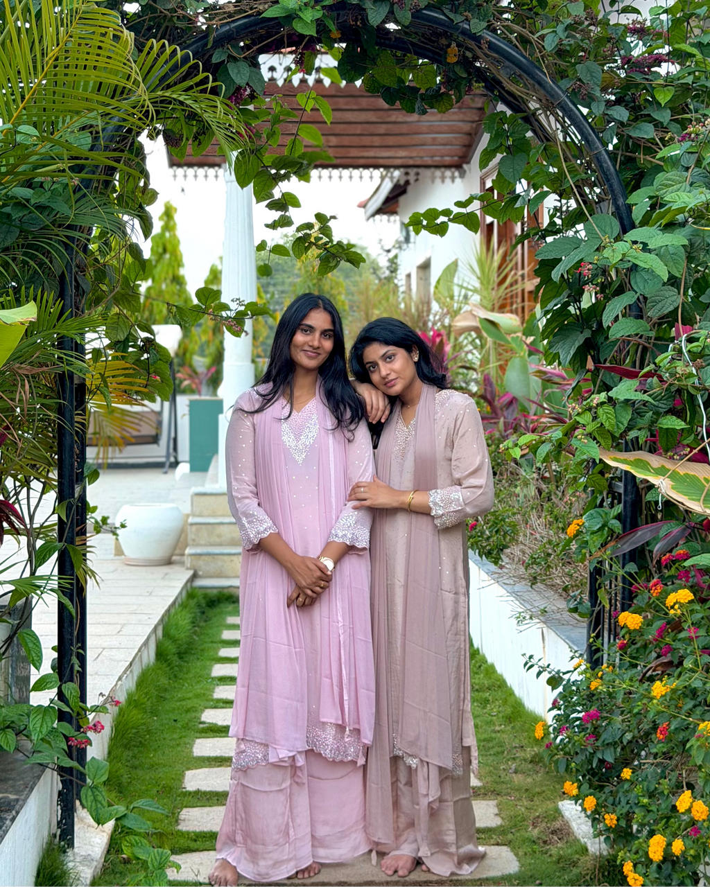 Two women in pink dresses standing under a floral archway in a garden.