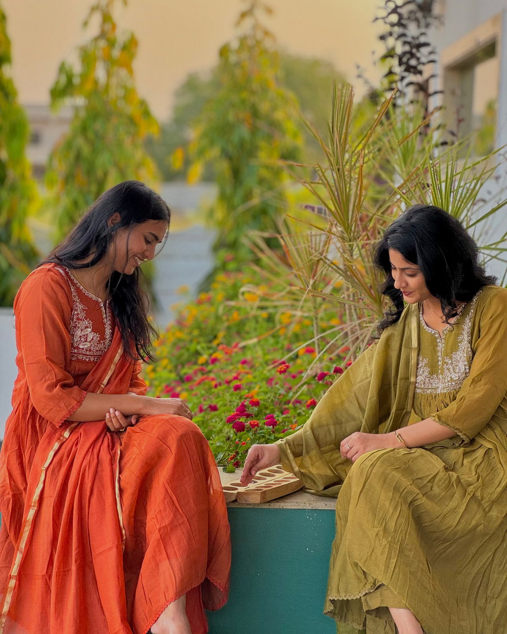 Two women in traditional attire sitting on a ledge with a garden and lake in the background.