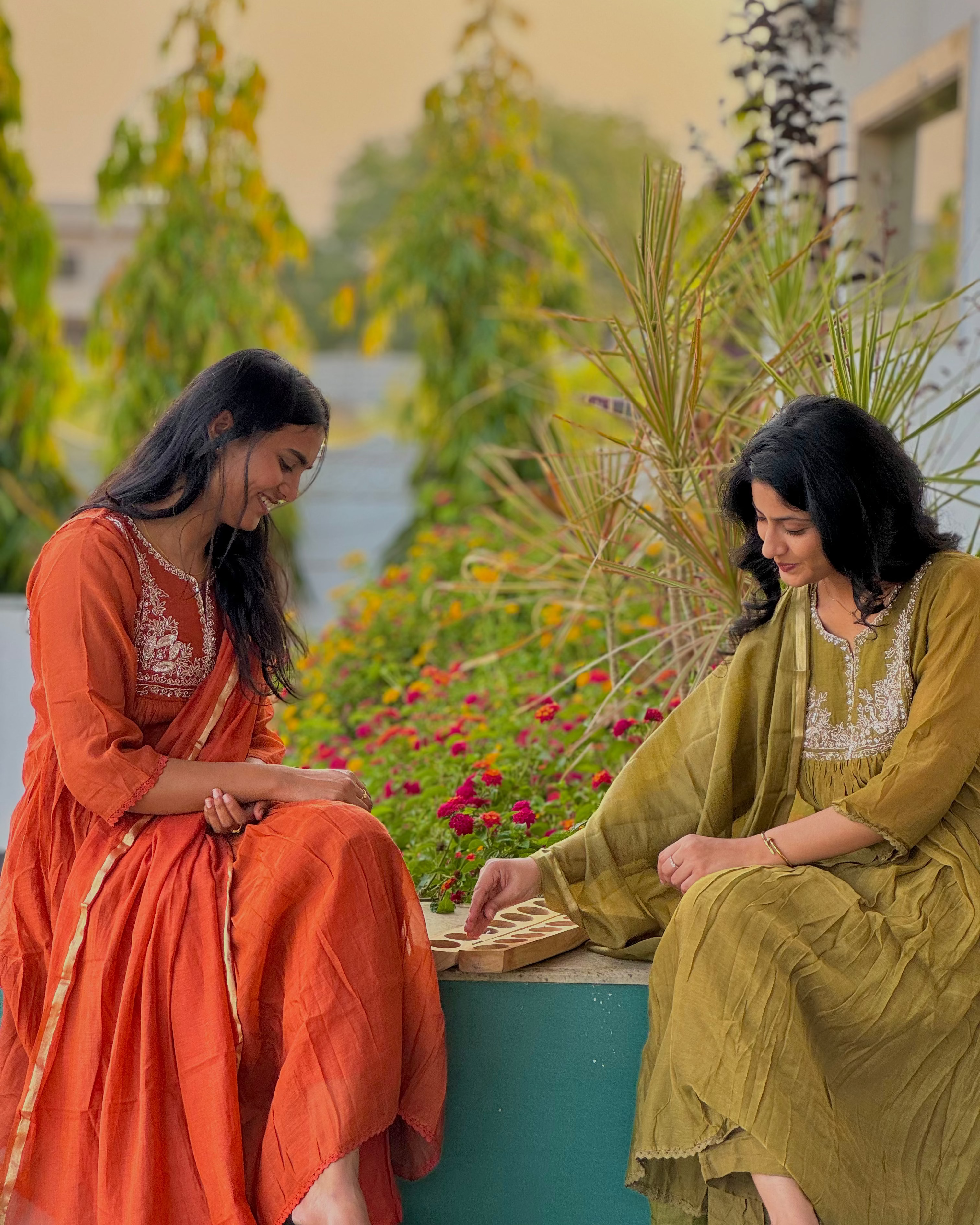 Two women in traditional attire sitting on a ledge with a garden and lake in the background.