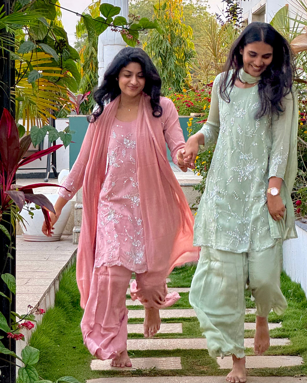 Two women in traditional outfits walking outdoors on a wooden path with greenery.
