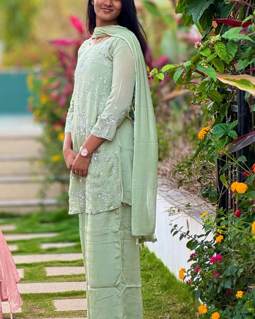 Woman in a light green traditional outfit standing in a garden with flowers and plants.