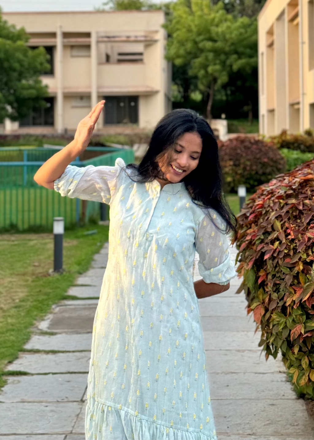 Woman in a light blue dress waving in front of a building with greenery