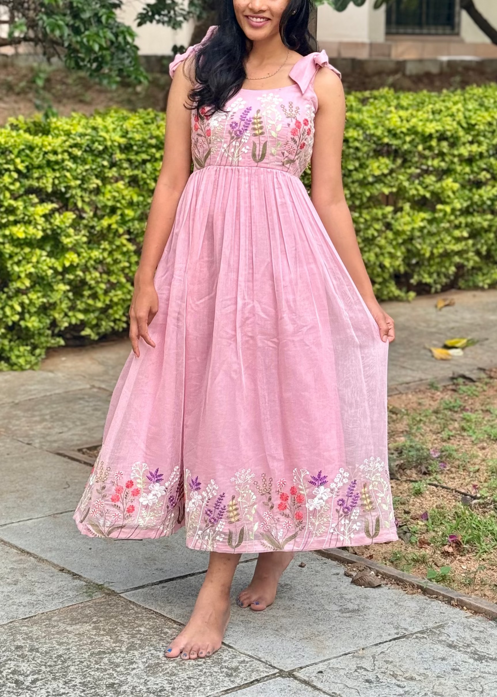 Woman in a pink dress standing outdoors with greenery and a building in the background