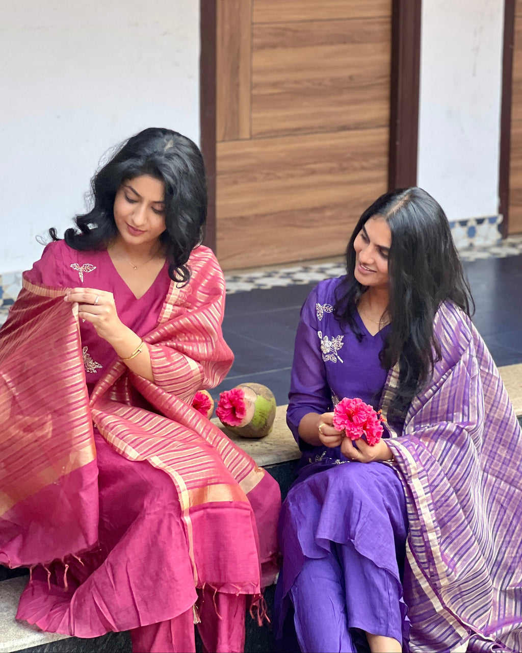 Two women sitting on a step, one in a pink saree and the other in a purple saree, holding flowers.