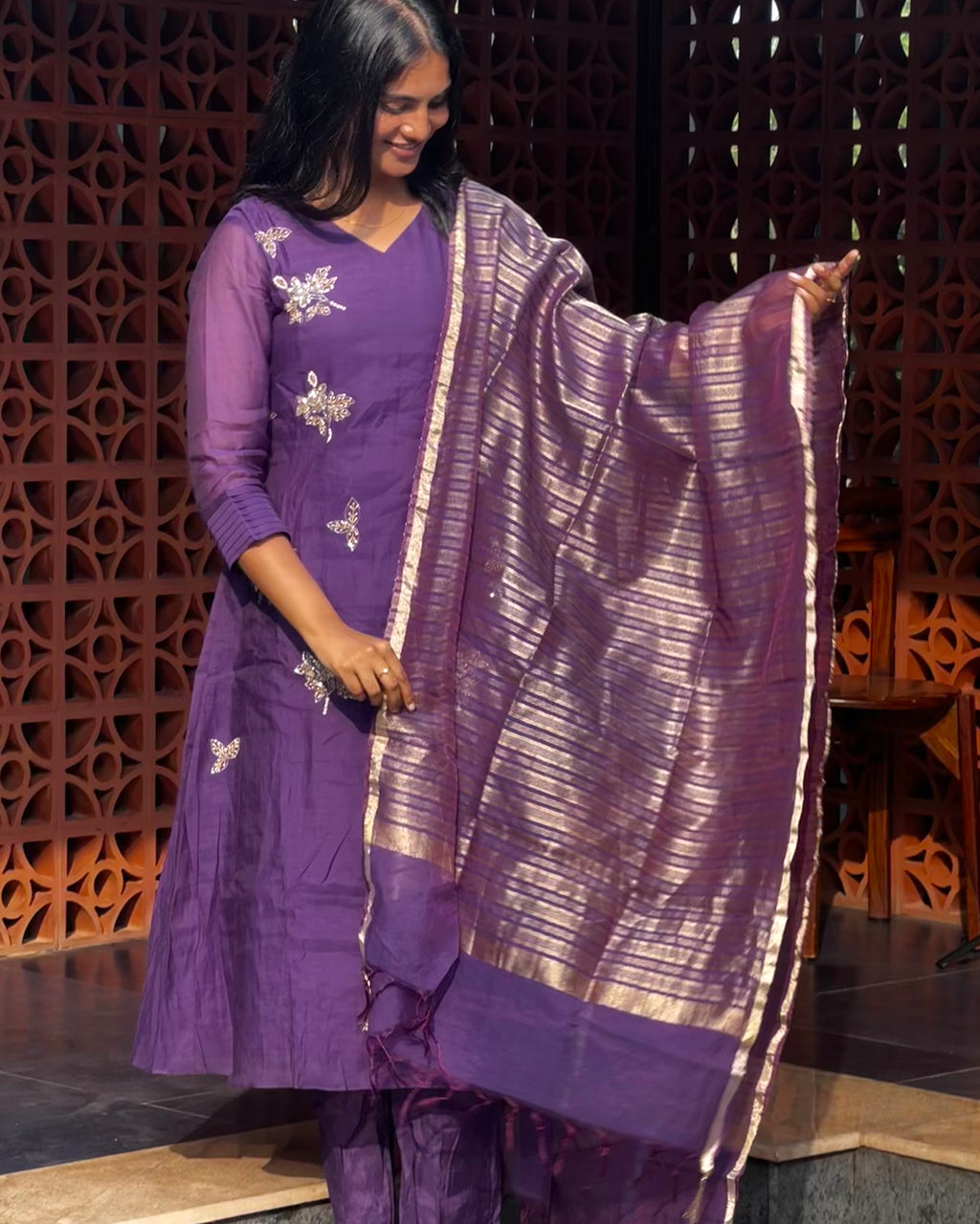Woman in a purple traditional outfit with a matching dupatta against a decorative wall.